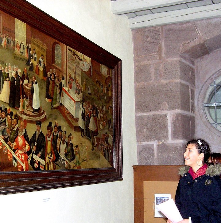 Fotoaufnahme der Führung „Bekenntnis – wozu wir uns bekennen“ in der Bad Windsheimer Spitalkirche. An einer Wand hängt ein Gemälde im Holzrahmen. Davor stehen zwei Jugendliche, die es betrachten. Die Wand rechts daneben besteht aus Sandsteinquadern. 