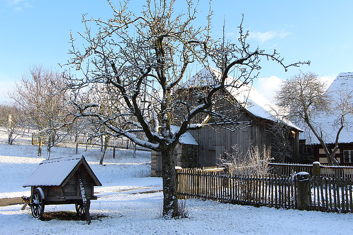 Fotoaufnahme der Scheune aus Dörflein und des Köblerhauses aus Oberfelden am aktuellen Standort. Auf beiden Gebäuden und am Boden liegt Schnee. Im Vordergrund steht ein kahler Baum, daneben steht ein kleiner überdachter Wagen aus Holz, dessen Dach ebenfalls schneebedeckt ist. 
