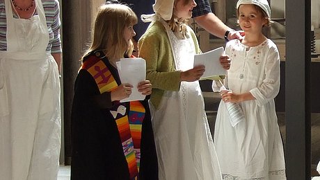Fotoaufnahme einer Szene aus dem Theaterstück zum Thema Gründung und Leben eines mittelalterlichen Spitals. Im Zentrum stehen drei kostümierte Mädchen mit Textblättern in der Hand. Dahinter sind zwei Erwachsene, die das Schauspiel beobachten.