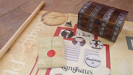 Fotoaufnahme der Utensilien zur Schatzsuche im Museum Kirche in Franken. Auf dem Steinboden befindet sich der aufgerollte Schatzplan, darauf liegen ein versiegelter Schatzbrief und eine kleine Schatzkiste aus Holz. 