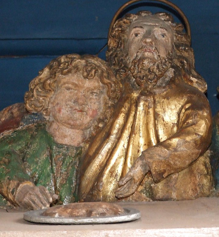 Fotoaufnahme einer figürlichen Darstellung des Abendmahls. Bei der Figur rechts mit Bart und Heiligenschein handelt es wohl um Jesus. Er gestikuliert mit seiner Hand in Richtung des Brotes, das auf dem Tisch vor ihm liegt. Die Figur links ist kleiner und lehnt sich an Christus an. 